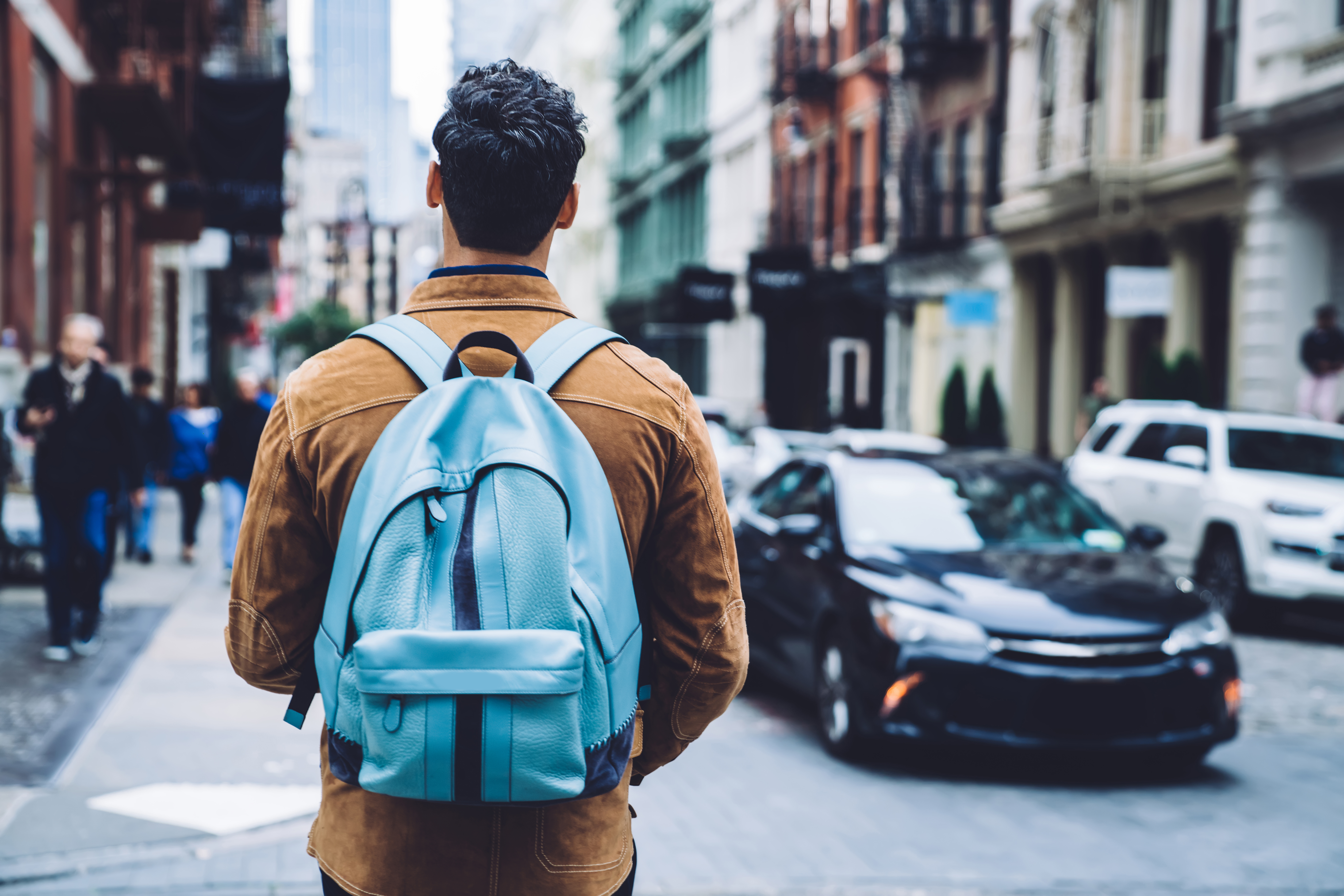 Vue arrière d’un jeune homme portant une veste marron et un sac à dos bleu marchant dans une rue, en ville